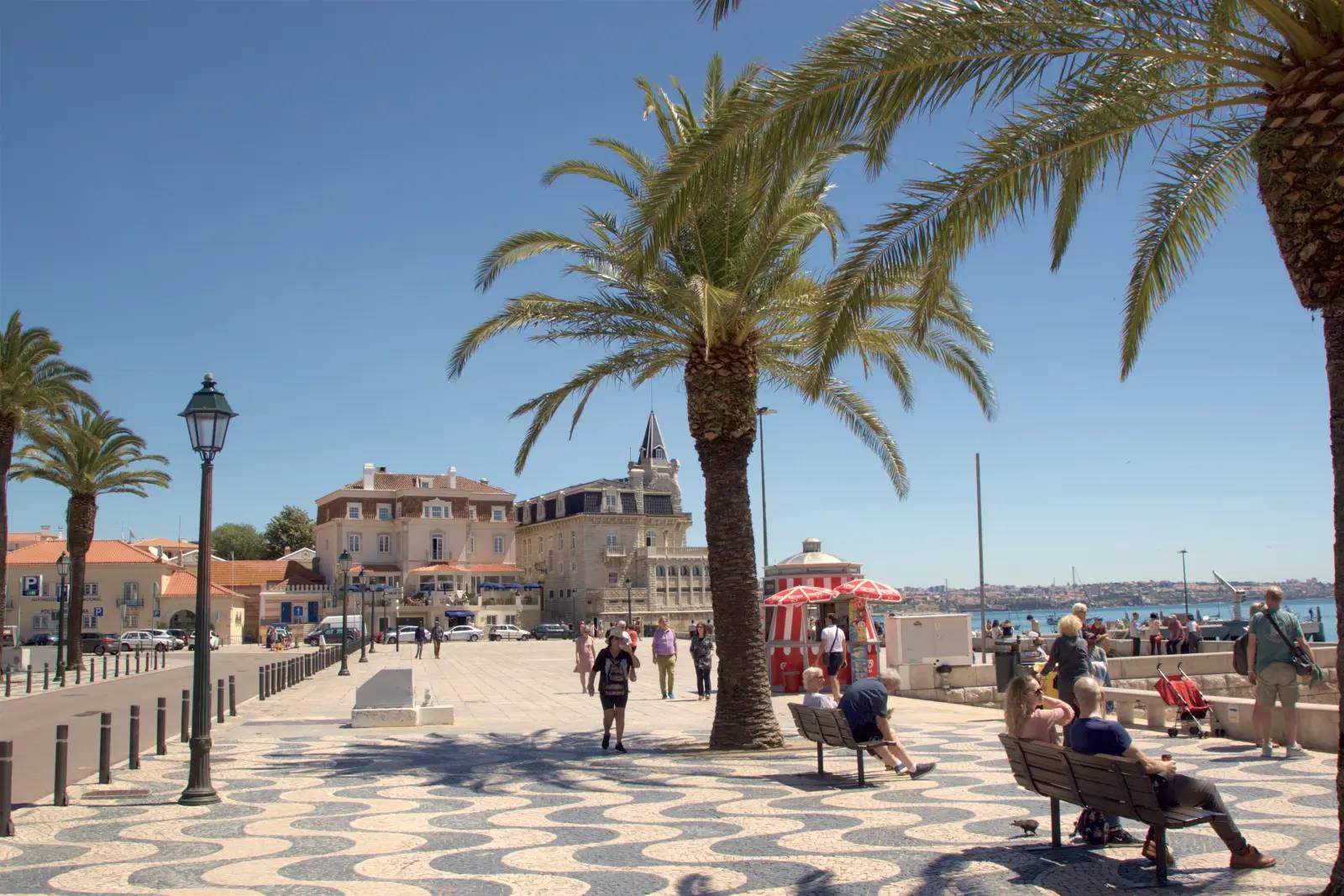 People relaxing on benches under palm trees on the seafront promenade of Cascais, near Lisbon, on a sunny day