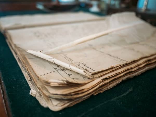 Um antigo livro de registo de contas aberto, com lançamentos manuscritos e uma pena de escrever sobre uma secretária verde