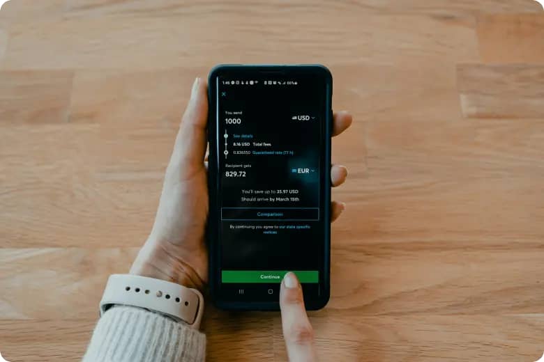 Main tenant un téléphone affichant le devis d’une application de transfert d’argent : 1,000 USD envoyés, 829.72 EUR reçus