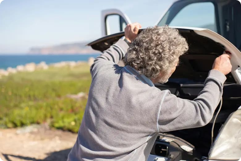 Man die de motorkap optilt van een witte auto die aan zee geparkeerd staat